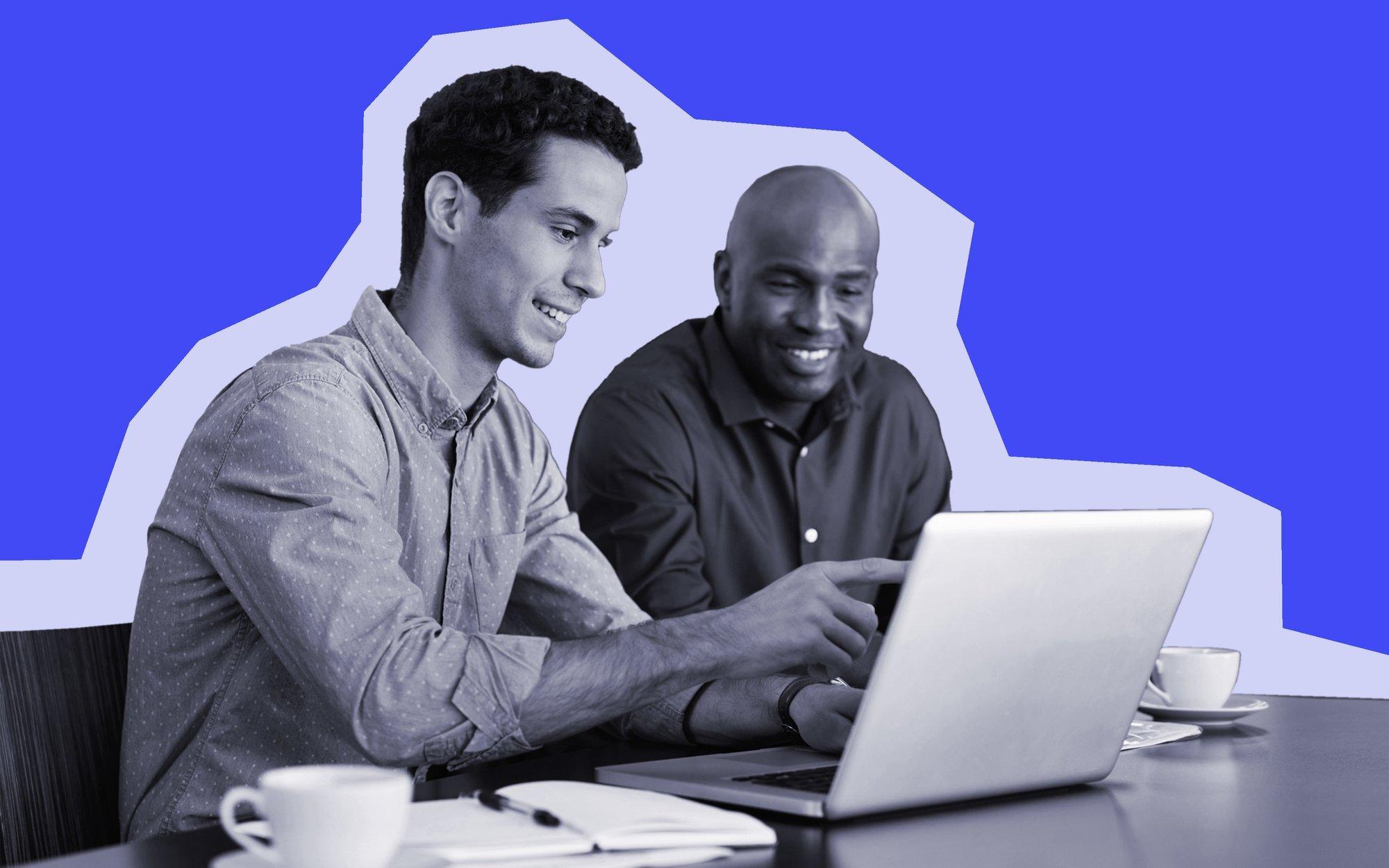 Two coworkers are sitting at a desk and smiling. One has a laptop open and is pointing at the screen.